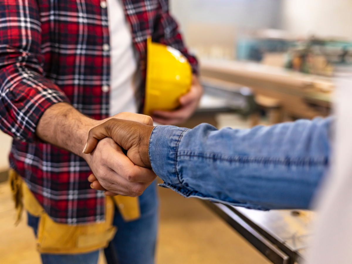 Close-up of two people shaking hands, one holding a yellow hard hat, symbolizing a professional agreement or partnership in a construction setting.