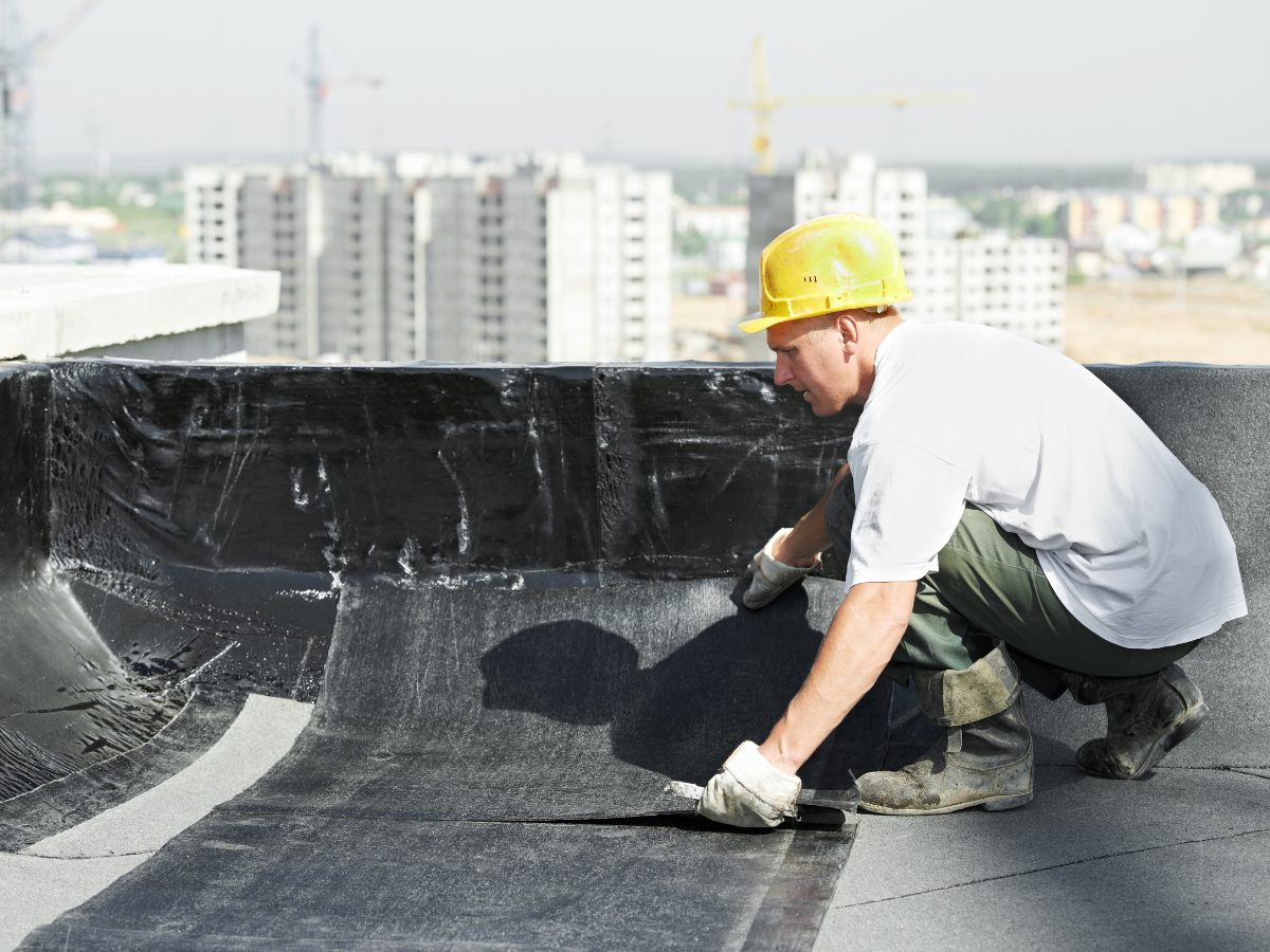 Construction worker in a yellow hard hat installing roofing material on a flat commercial roof, with a city skyline in the background.