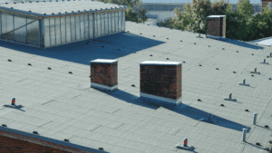 Flat commercial rooftop with two brick chimneys and a bitumen roofing system, showing smooth gray surface with vent pipes and equipment mounts across the roof.