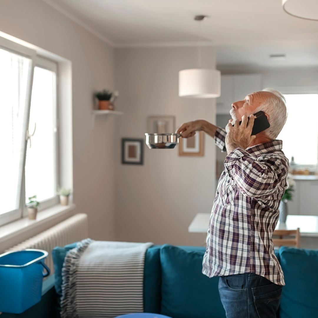 Older man holds a pan to catch a drip from a leak in his ceiling.