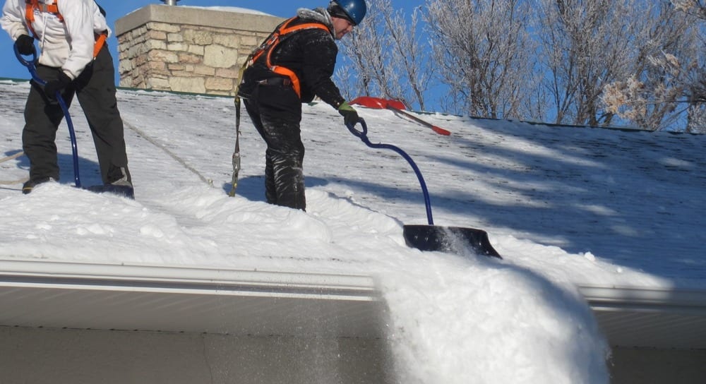 Roofers wearing orange harnesses and hard hats. They're dressed in clothing to keep them warm in the snow. They are standing on the roof of a home shoveling snow off the roof.