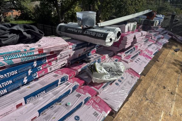 Roofing materials and supplies stacked across a residential rooftop during an active roofing project, including wrapped shingles, underlayment, tools, and metal flashing under bright sunlight