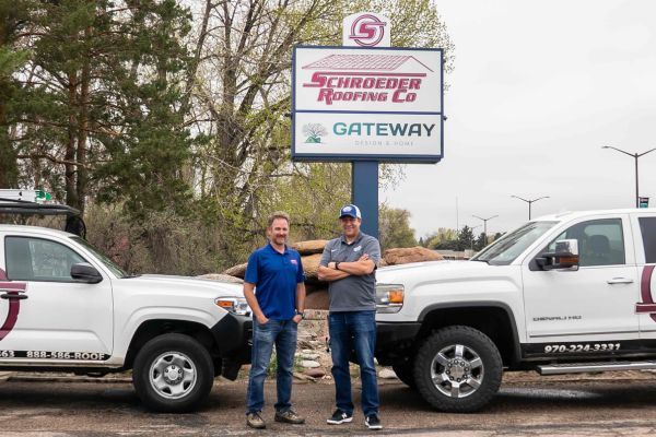 Two men stand between two white company trucks branded with the Schroeder Roofing Co logo in front of a large roadside sign for Schroeder Roofing Co and Gateway Design & Home. The men pose with arms relaxed and crossed, wearing casual work attire, in a parking lot lined with trees on an overcast day.
